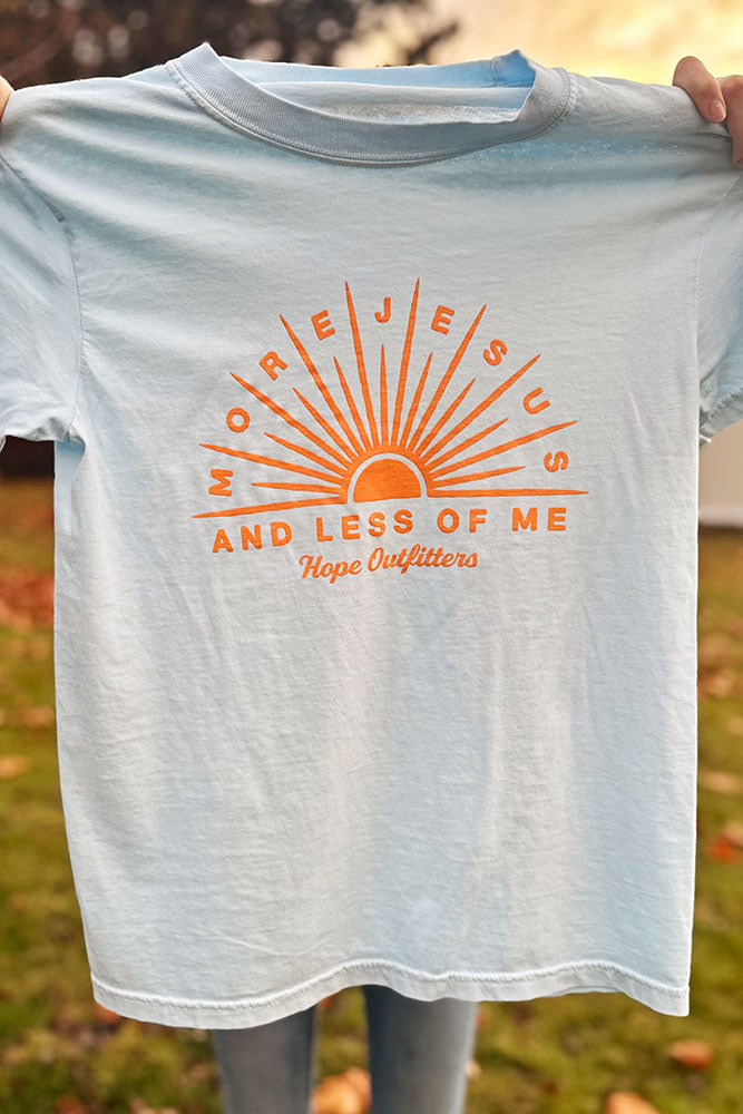 More Jesus & Less of Me Christian t-shirt, light blue with orange sunburst print, held up