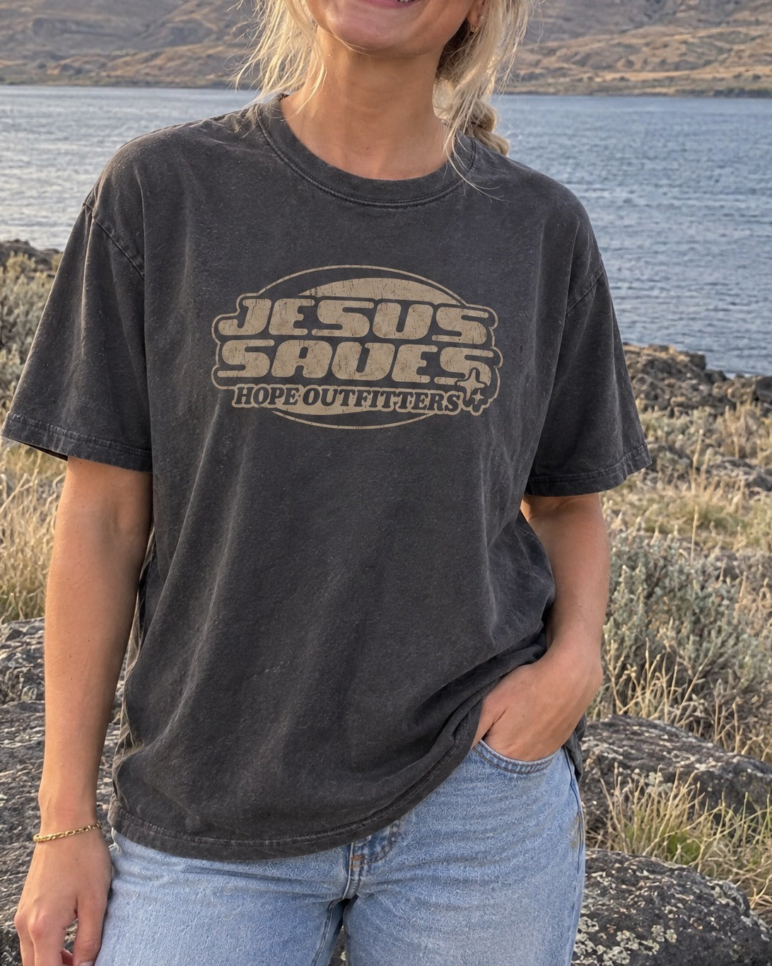 Jesus Saves Retro Tee - close-up of the graphite tee print worn by a blonde woman on basalt rocks