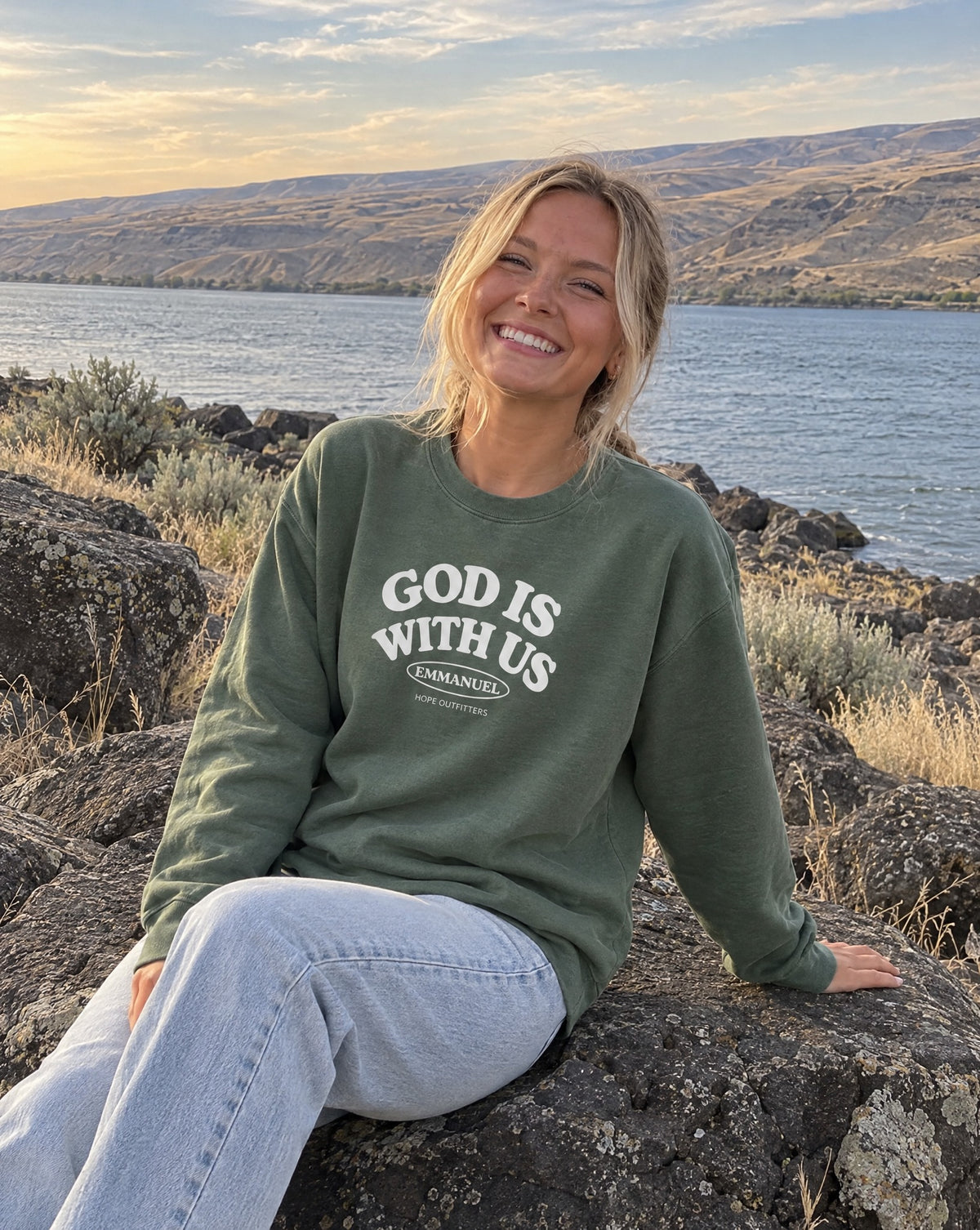Emmanuel God With Us Crewneck Sweatshirt - blonde woman sitting on a basalt boulder by the river in the Military Green crewneck