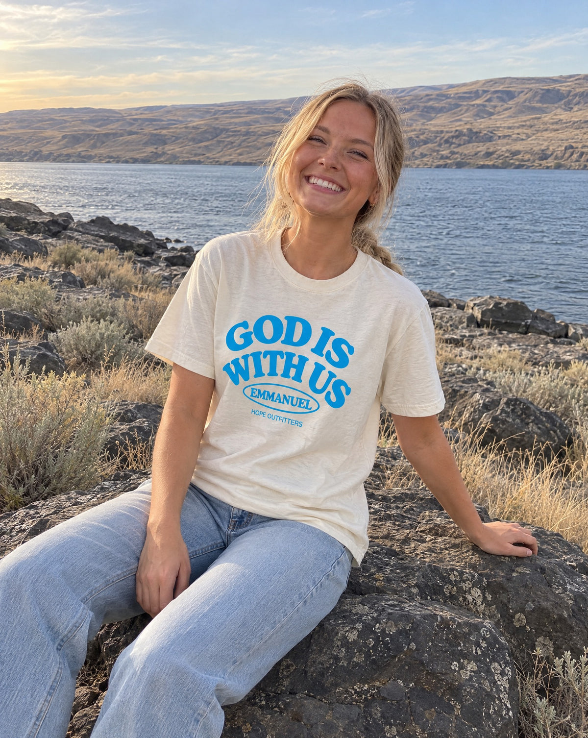 Emmanuel God With Us Tee - blonde woman sitting on a basalt boulder by the river in the natural tee