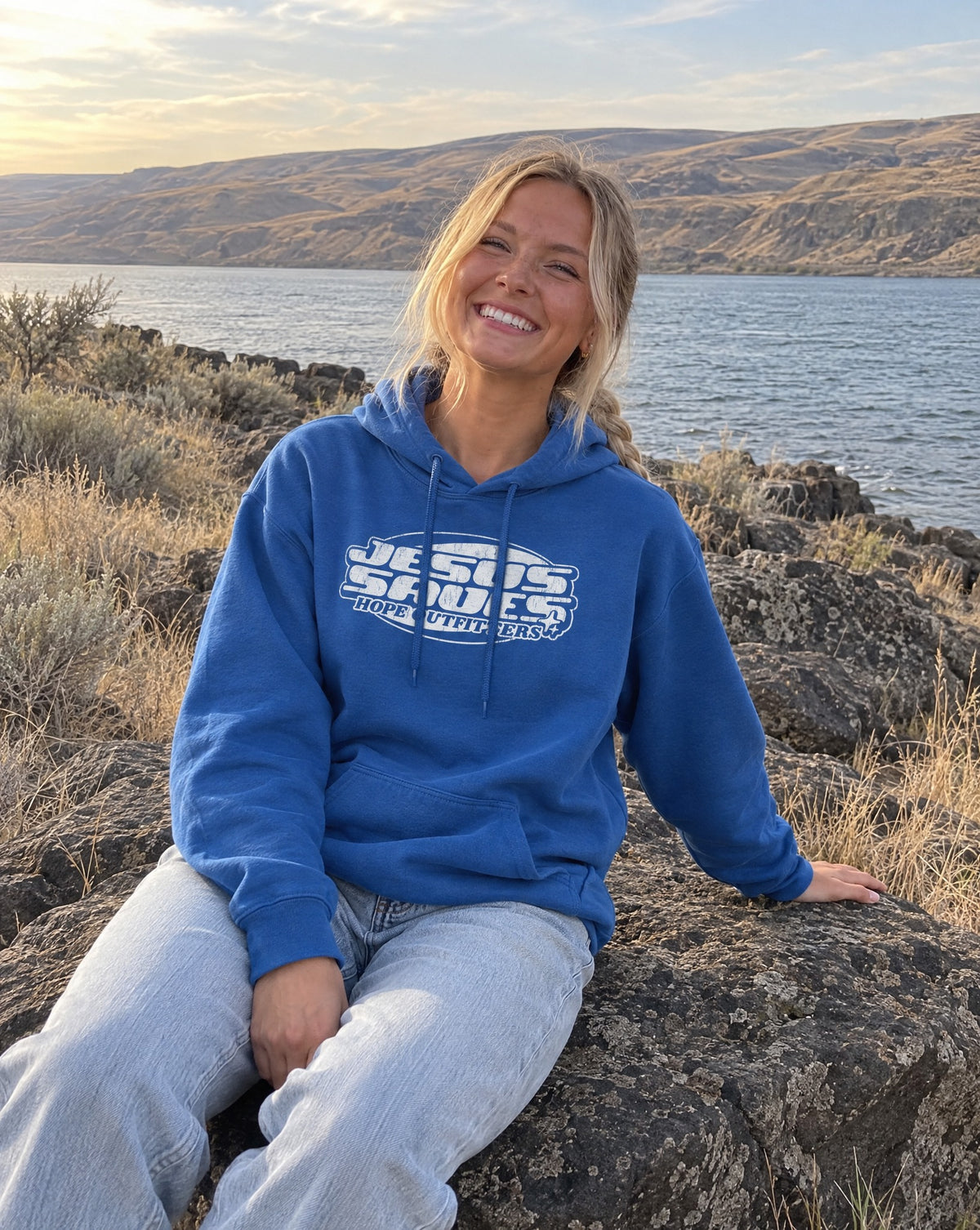 Jesus Saves Retro Hoodie - blonde woman sitting on a basalt boulder by the river in the Royal blue hoodie