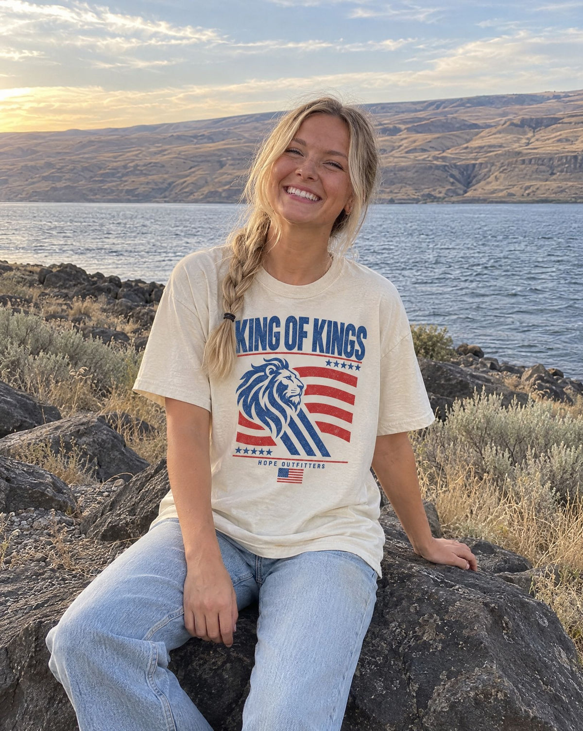 King of Kings Lion Tee - blonde woman sitting on a basalt boulder by the river in the ivory tee