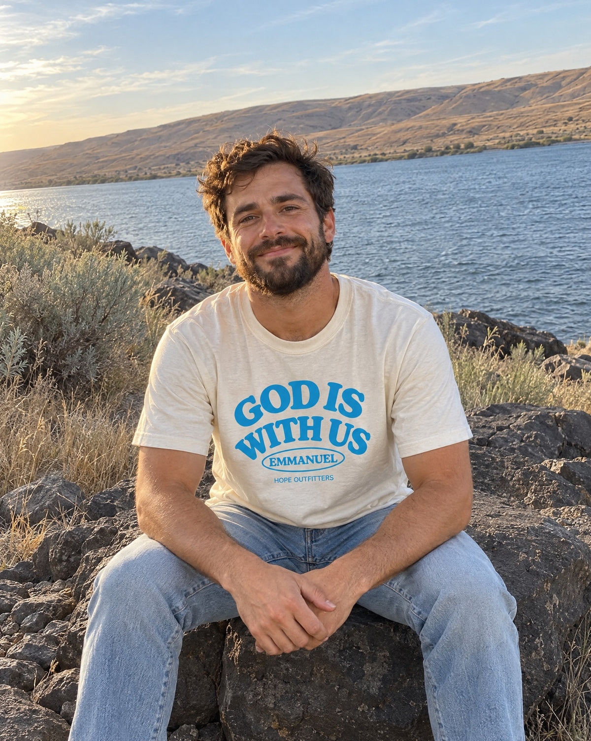 Emmanuel God With Us Tee - bearded man sitting on basalt by the river in the natural tee