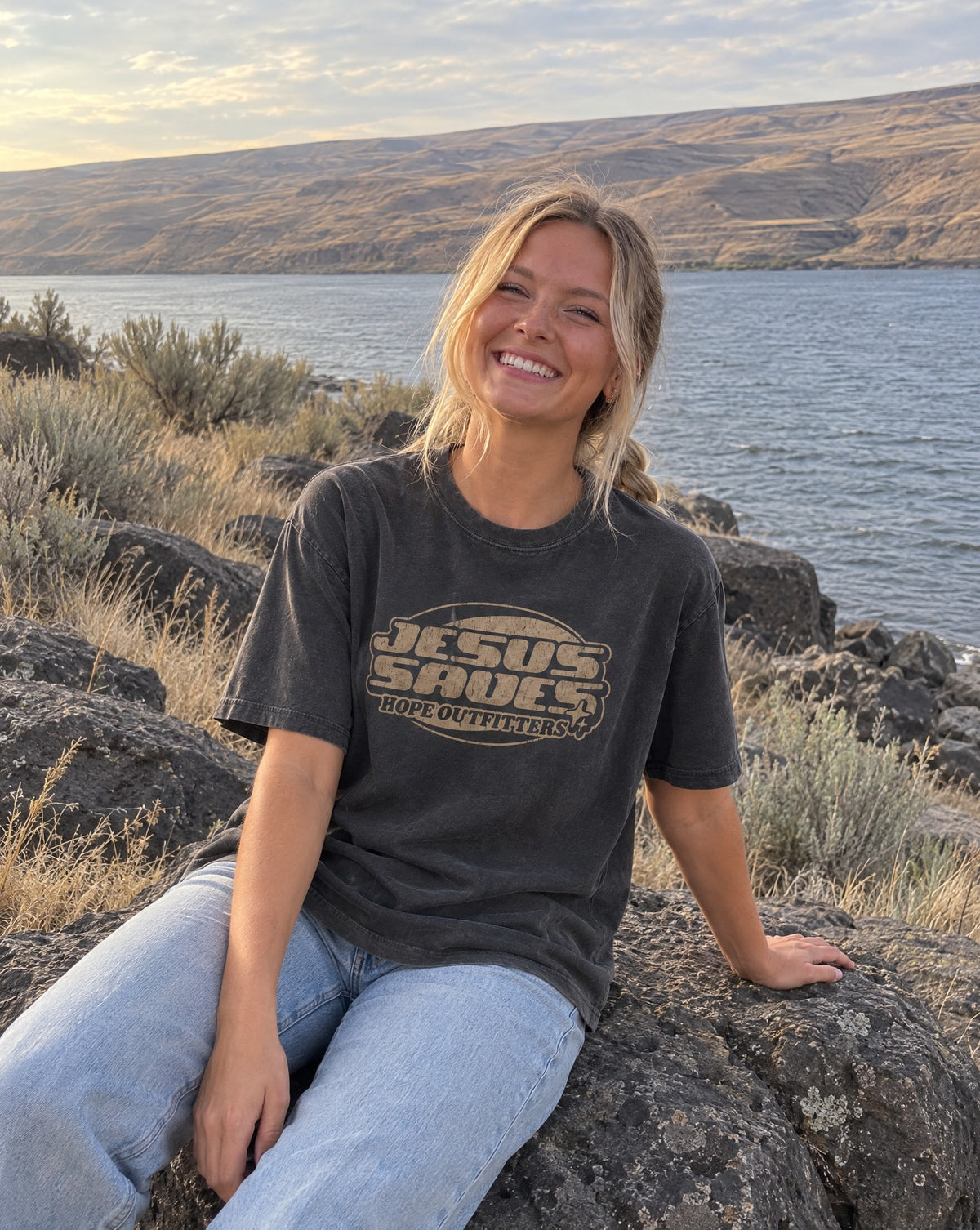 Jesus Saves Retro Tee - blonde woman sitting on a basalt boulder by the river in the graphite tee