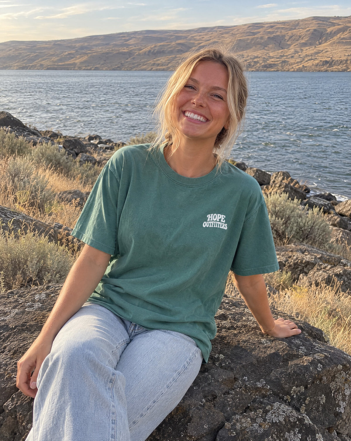 No Other Name But Jesus Tee - blonde woman sitting on a basalt boulder by the river in the emerald tee
