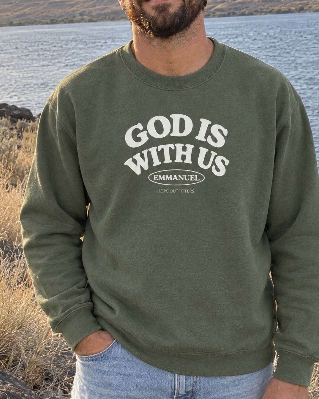 Emmanuel God With Us Crewneck Sweatshirt - close-up of the Military Green crewneck's white God Is With Us print worn by a bearded man by the Columbia River