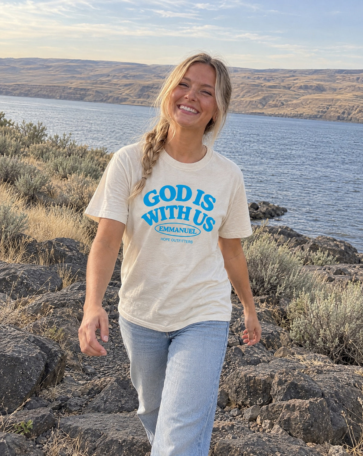 Emmanuel God With Us Tee - blonde woman walking over basalt rocks by the Columbia River in the natural tee