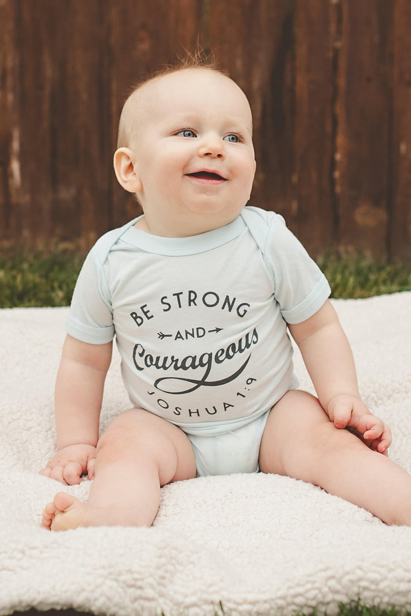 Baby sitting up wearing the white Be Strong and Courageous onesie